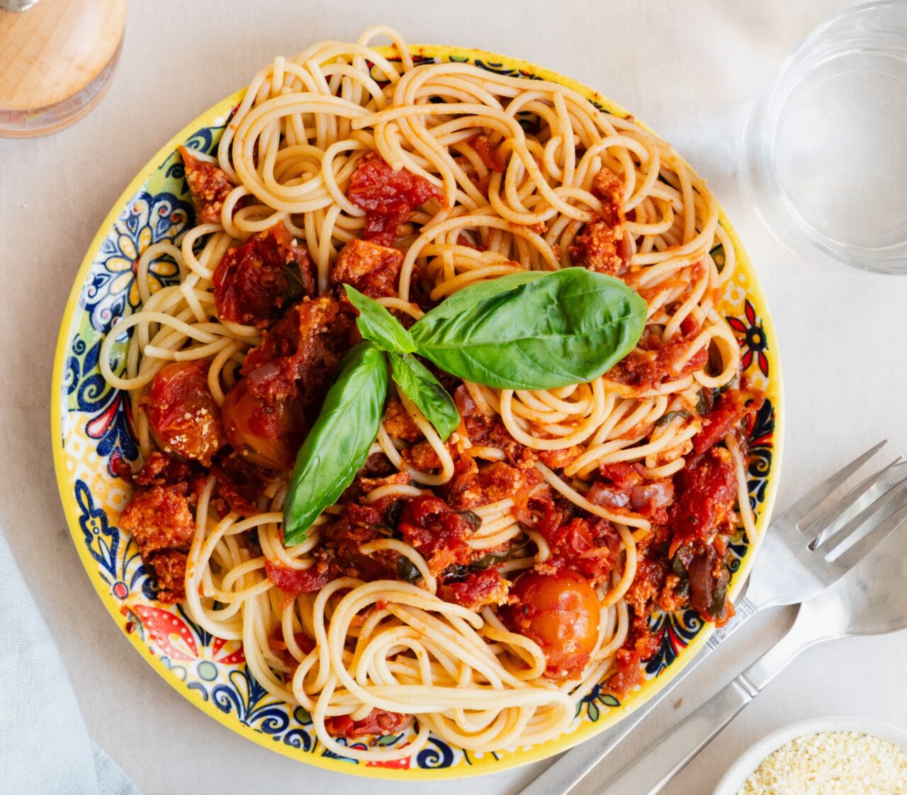 Easy Meatless Spaghetti Bolognese with crumbled tofu, tomatoes, and fresh basil on a colorful patterned plate, with a fork and a small bowl of dairy-free parmesan on the side.