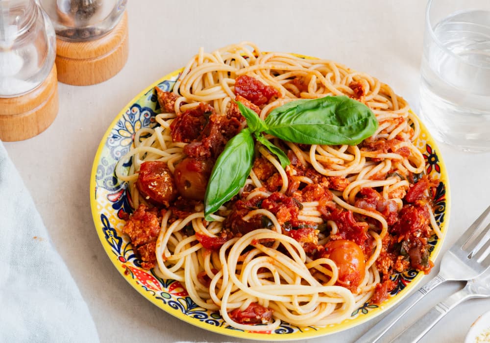 Plate of meatless spaghetti bolognese with crumbled tofu, tomatoes, and fresh basil on a colorful patterned plate, next to wooden salt and pepper mills, a glass of water, and a fork.
