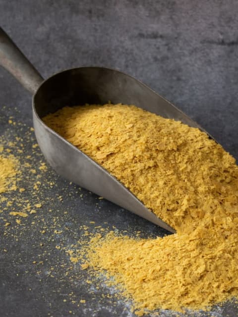 A metal scoop of golden nutritional yeast flakes spilling onto a dark slate surface.