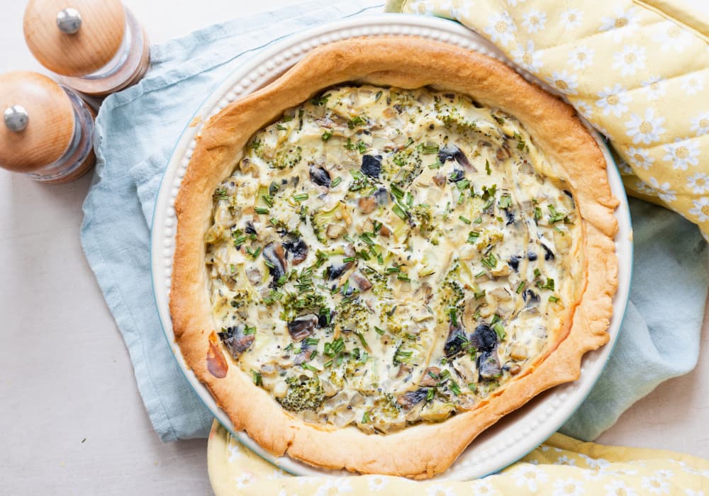 Overhead view of a whole eggless tofu quiche topped with fresh chives, in a white pie dish on a light blue napkin with yellow daisy oven mitts and wooden salt and pepper mills.