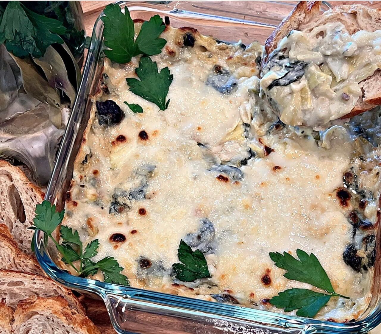 Baked Dairy-Free Spinach Artichoke Dip in a square glass baking dish with a bubbly, golden-browned top and fresh parsley, with a slice of toasted sourdough scooping out a bite.