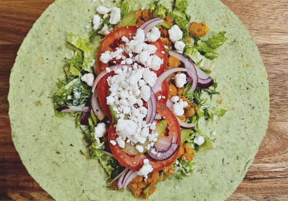 An open spinach tortilla layered with ranch-coated romaine, buffalo chickpeas, celery, sliced tomato, red onion, and crumbled vegan feta.