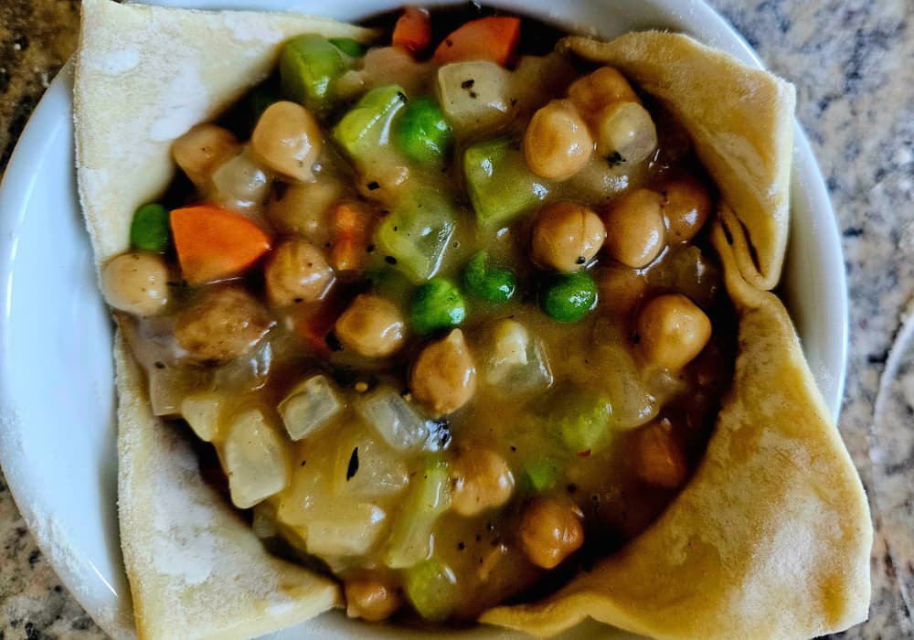 A pastry-lined ramekin filled with chickpeas, carrots, celery, peas, and onions in a savory gravy, ready to be topped with puff pastry.