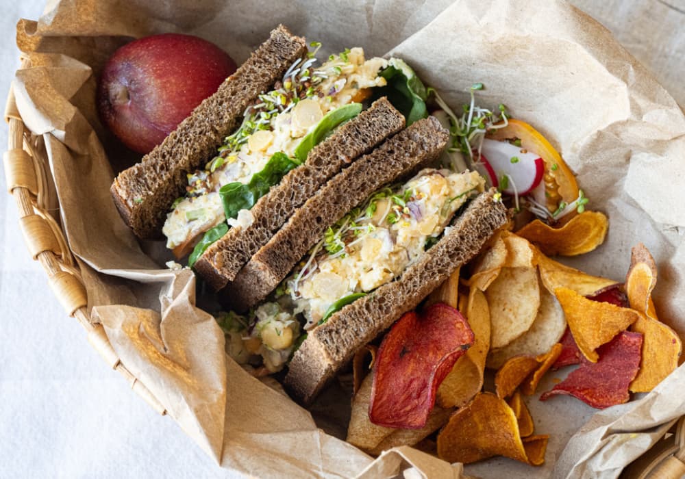 Chickpea Salad Sandwich on dark rye bread, cut in half and packed in a paper-lined basket with vegetable chips, sliced radish, and a red apple.
