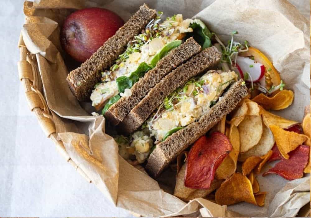 Chickpea Salad Sandwich on dark rye bread, cut in half and packed in a paper-lined basket with vegetable chips, sliced radish, and a red apple.
