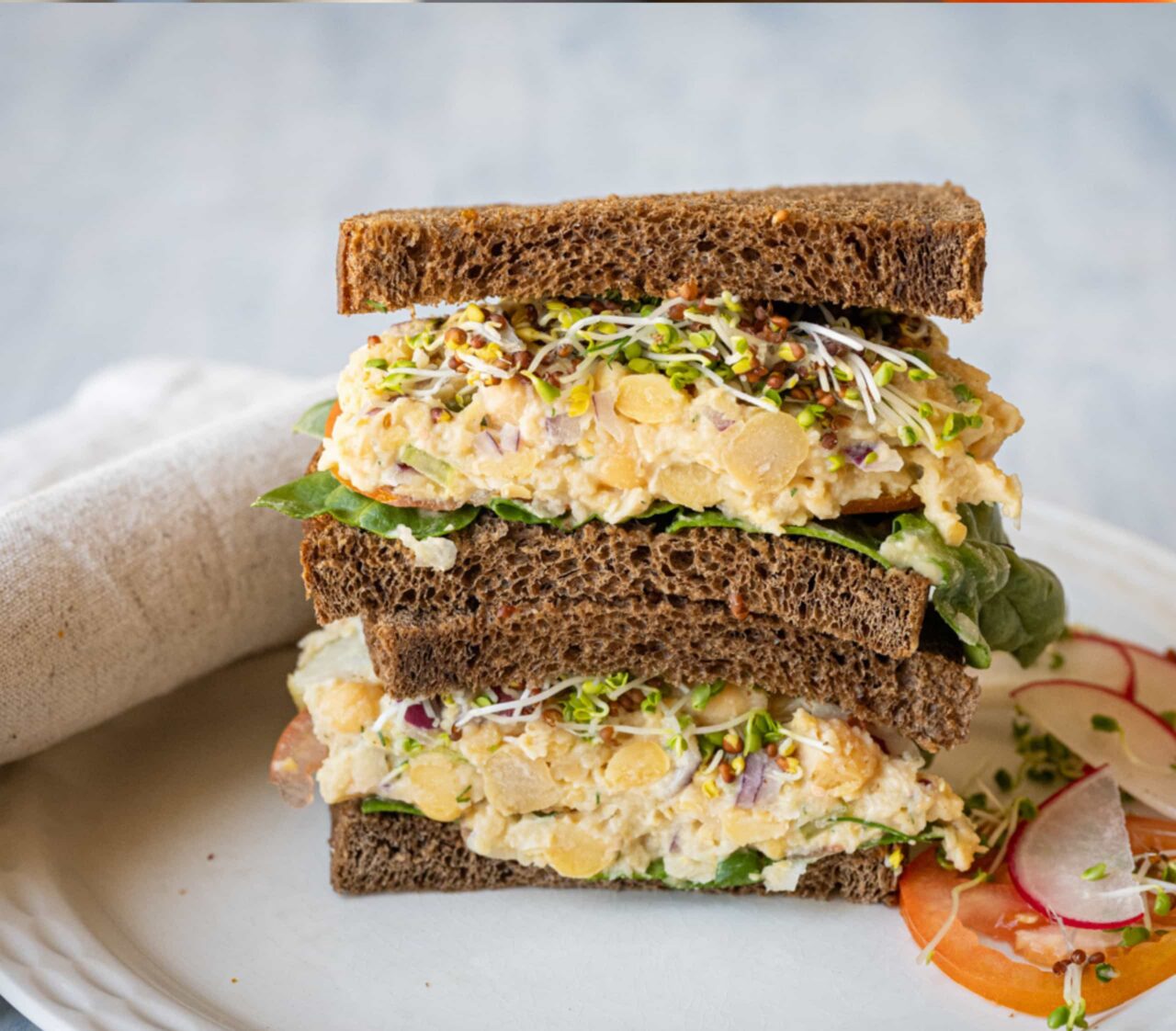 Chickpea Salad Sandwich on dark rye bread, cut in half and stacked on a white plate with spinach, sprouts, sliced radish, and tomato.