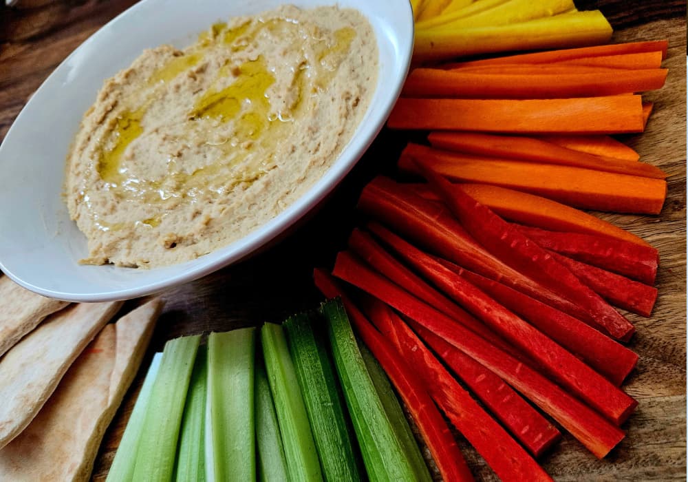 Angled close-up of Classic Homemade Hummus drizzled with olive oil, served with rainbow carrot sticks, celery, and pita wedges.
