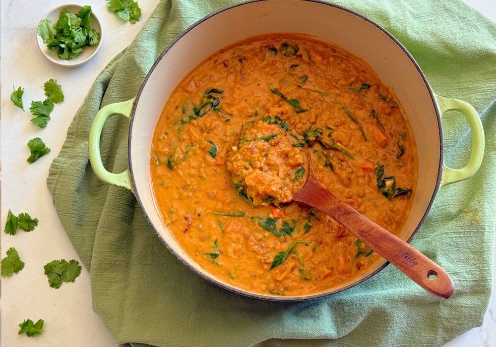 A pot of Coconut Lentil Soup with Butternut Squash and Spinach with a wooden ladle, set on a green kitchen towel with fresh cilantro.