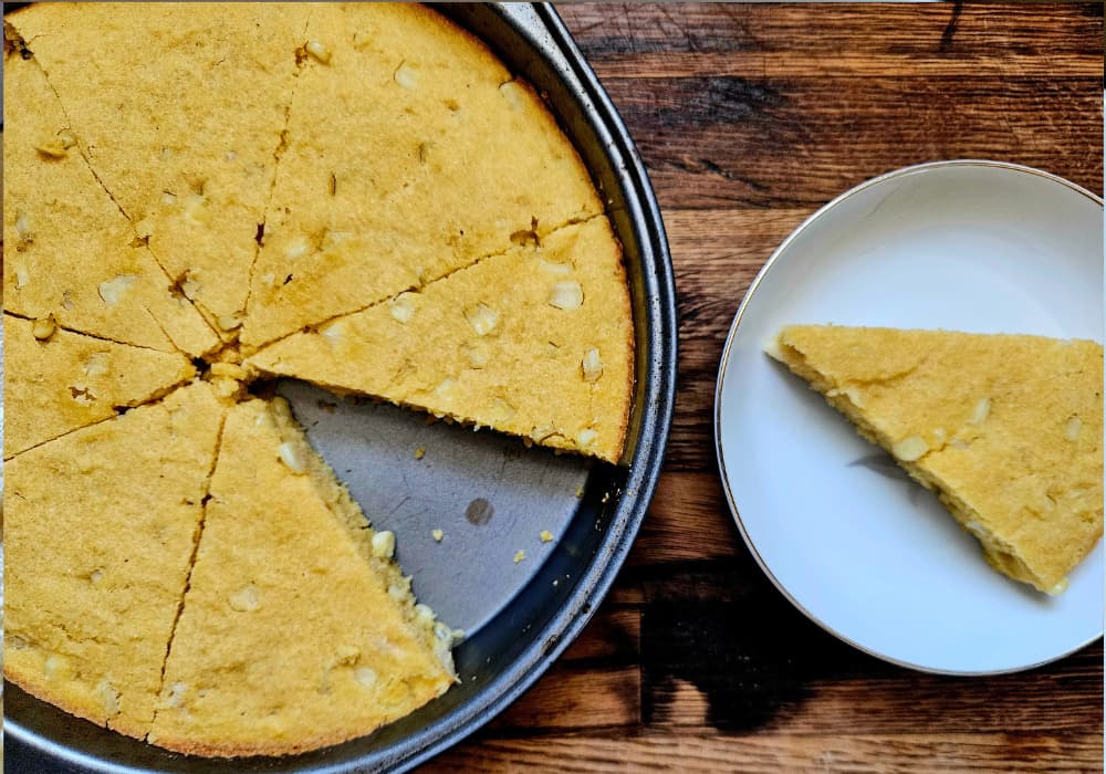 Sliced cornbread with sweet corn kernels in a round baking pan, with one wedge served on a white plate.