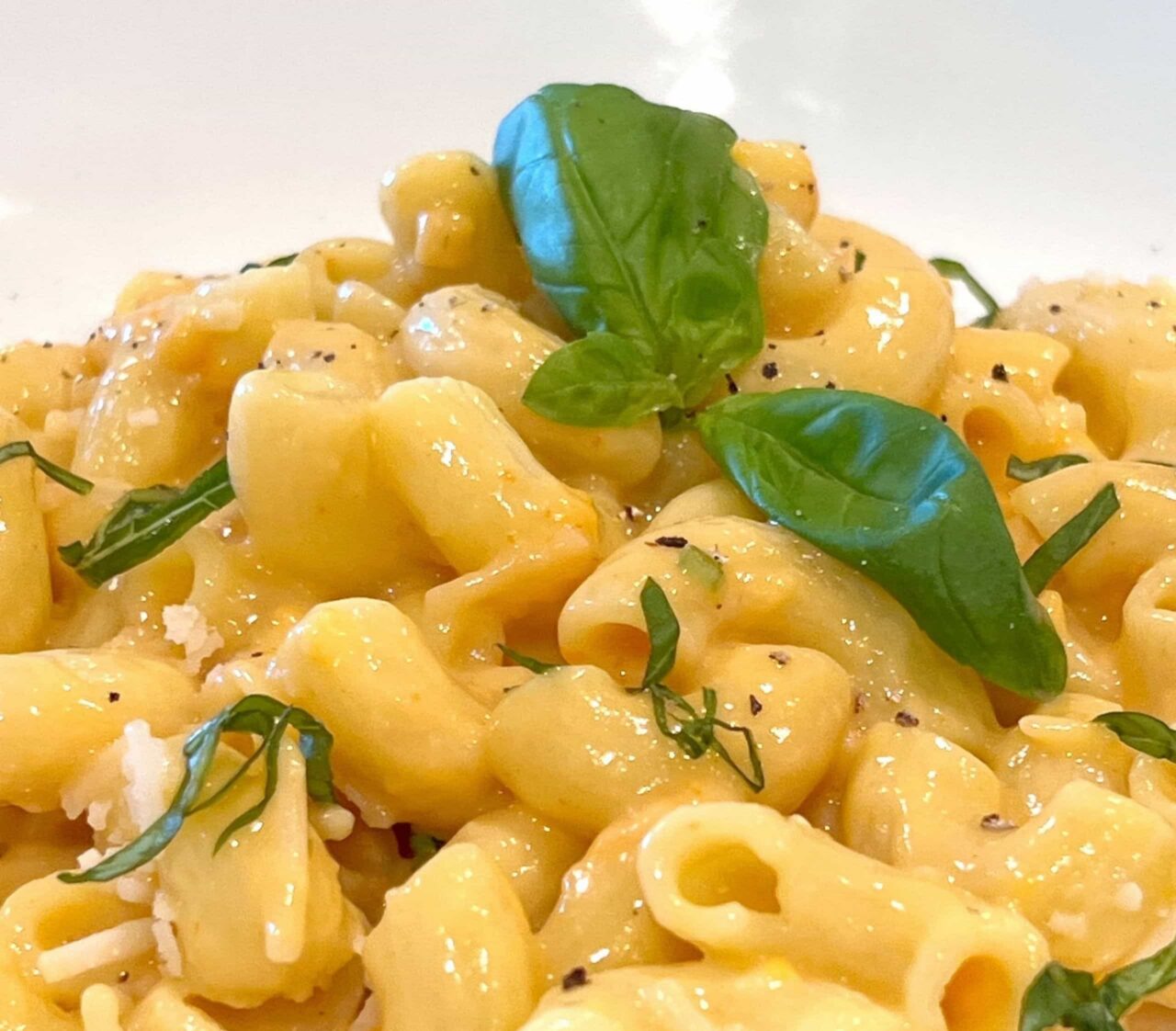 Close-up of Creamy Dairy-Free Mac and Cheese in a white bowl, with glossy cheese sauce, fresh basil leaves, grated dairy-free Parmesan, and black pepper.