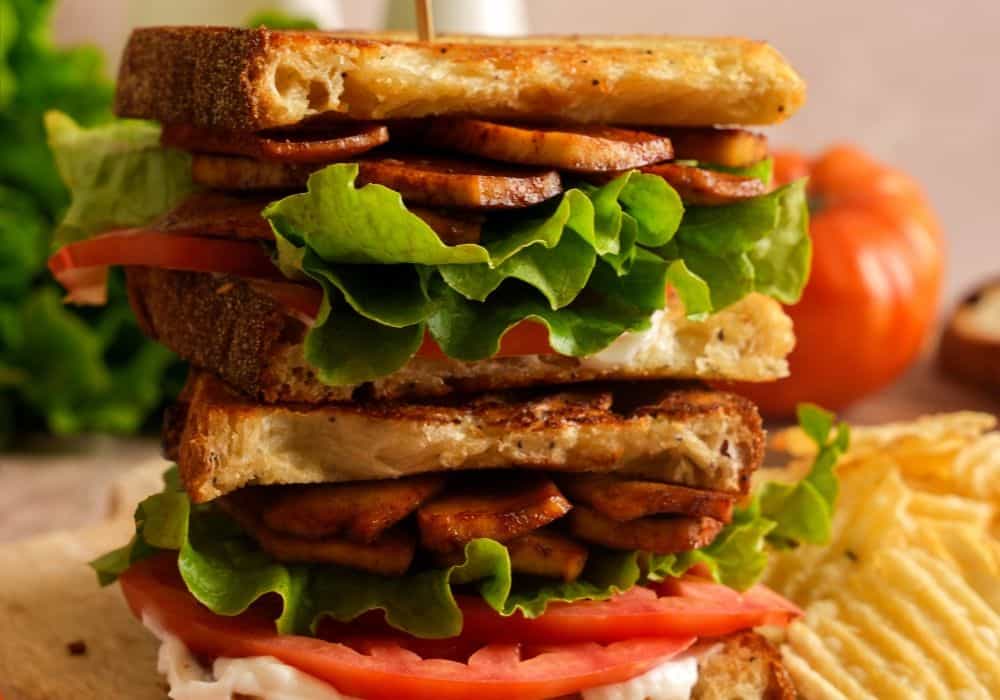Two halves of a crispy tofu BLT stacked on a wooden board with smoky tofu bacon, lettuce, tomato, and vegan mayo, served with potato chips.