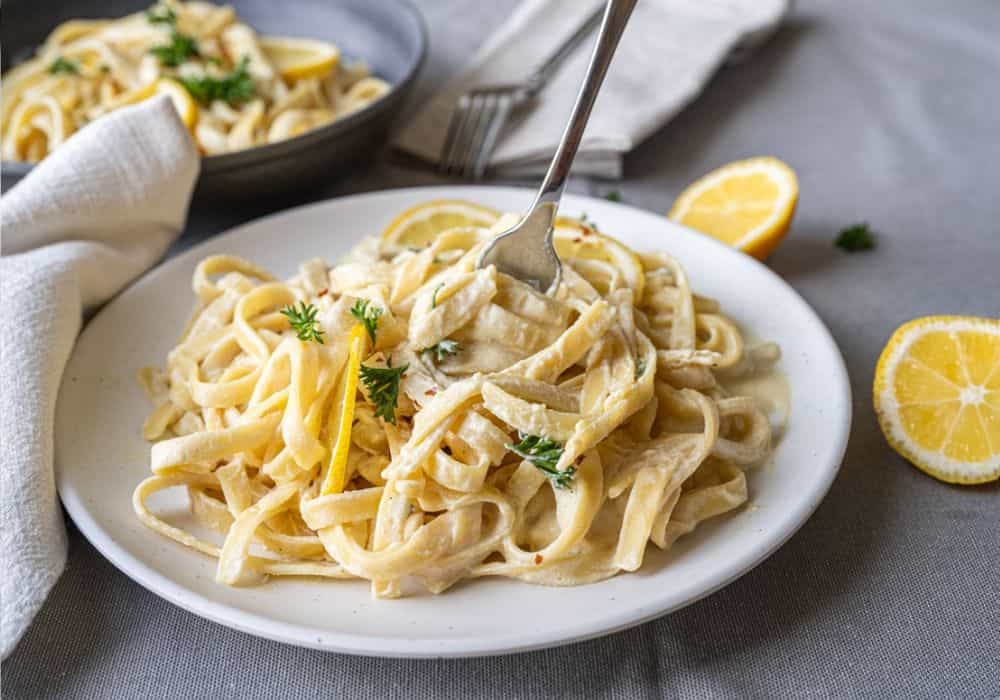 Fork lifting creamy dairy-free fettuccine Alfredo with silken tofu, garnished with parsley and lemon, on a white plate.