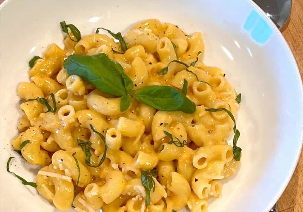 Overhead view of a white bowl of Creamy Dairy-Free Mac and Cheese topped with fresh basil, grated dairy-free Parmesan, and black pepper.