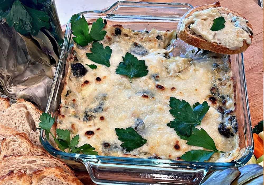 Baked Dairy-Free Spinach Artichoke Dip in a square glass baking dish topped with fresh parsley, with a slice of sourdough spread with dip, more sliced sourdough, and vegetables on a wooden board.
