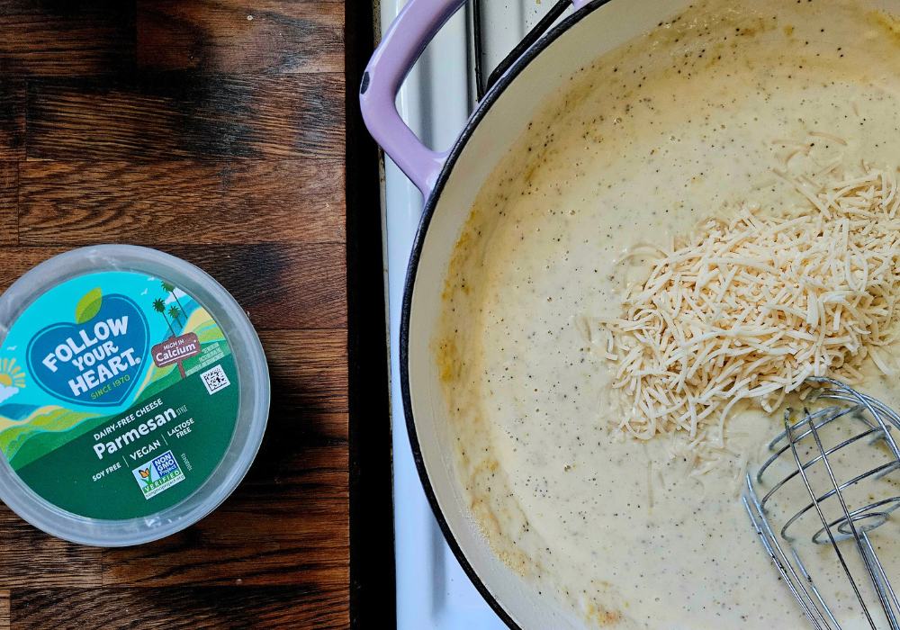 A container of Follow Your Heart dairy-free parmesan next to a pot of creamy sauce with shredded vegan parmesan being whisked in.