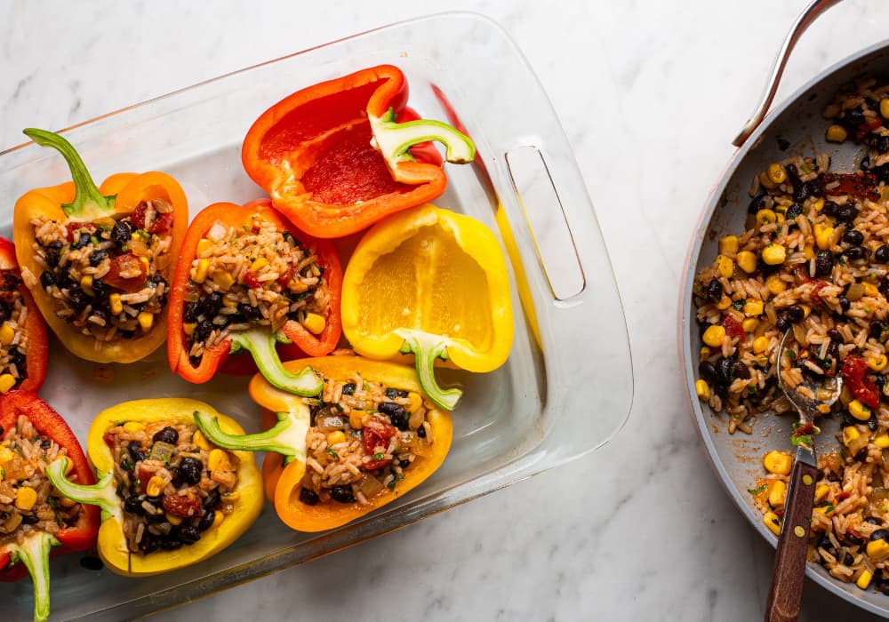 Red, orange, and yellow bell pepper halves being filled with a mixture of rice, black beans, corn, and tomatoes, next to a skillet of the filling.
