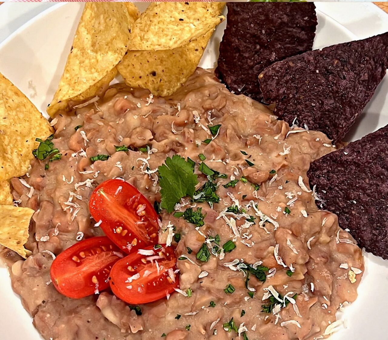 A white bowl of Instant Pot Refried Beans topped with grated dairy-free parmesan, chopped cilantro, and halved cherry tomatoes, with yellow and blue corn tortilla chips.