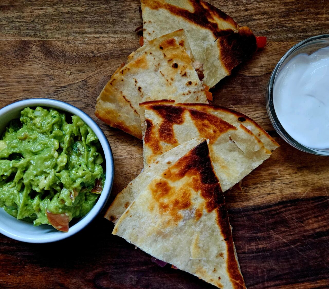 Golden, crispy Jackfruit Fajita Quesadillas cut into wedges on a wooden board, served with bowls of guacamole and sour cream.