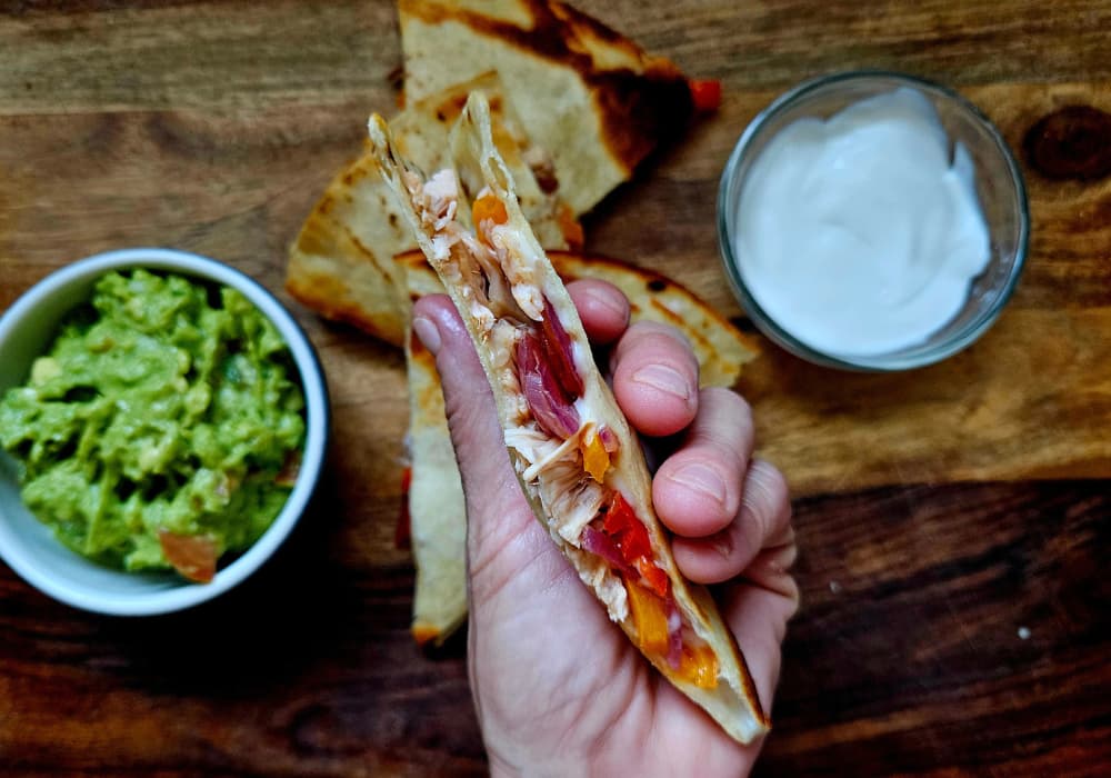 A hand holding a wedge of Jackfruit Fajita Quesadilla, showing the shredded jackfruit, peppers, onions, and melted cheese inside, with guacamole and sour cream on the side.