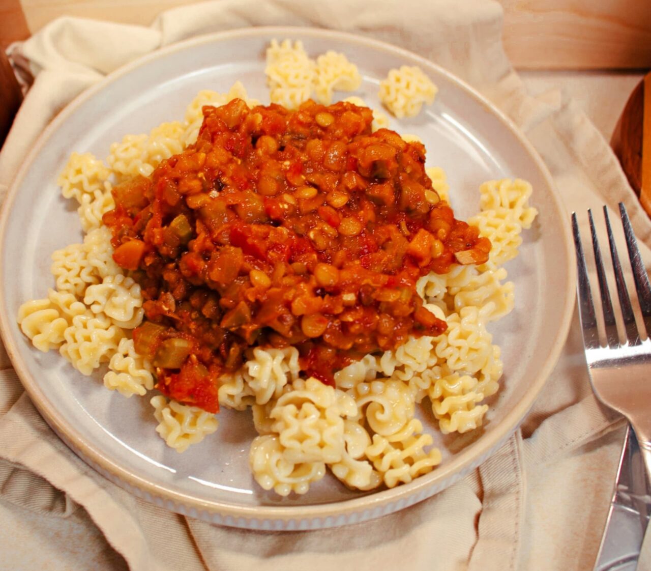 A plate of Hearty Lentil Bolognese spooned over pasta, with chunks of tomato, carrot, and lentils, served with a fork on a cream linen napkin.