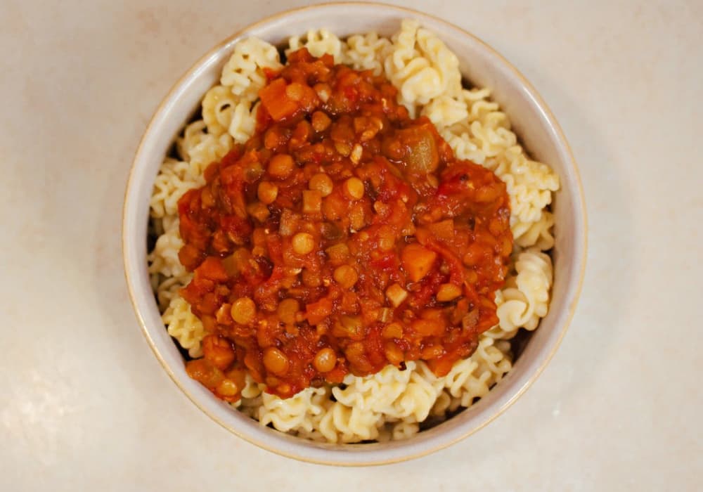 Overhead view of a bowl of pasta topped with Hearty Lentil Bolognese, showing red and brown lentils, diced carrots, and tomatoes.