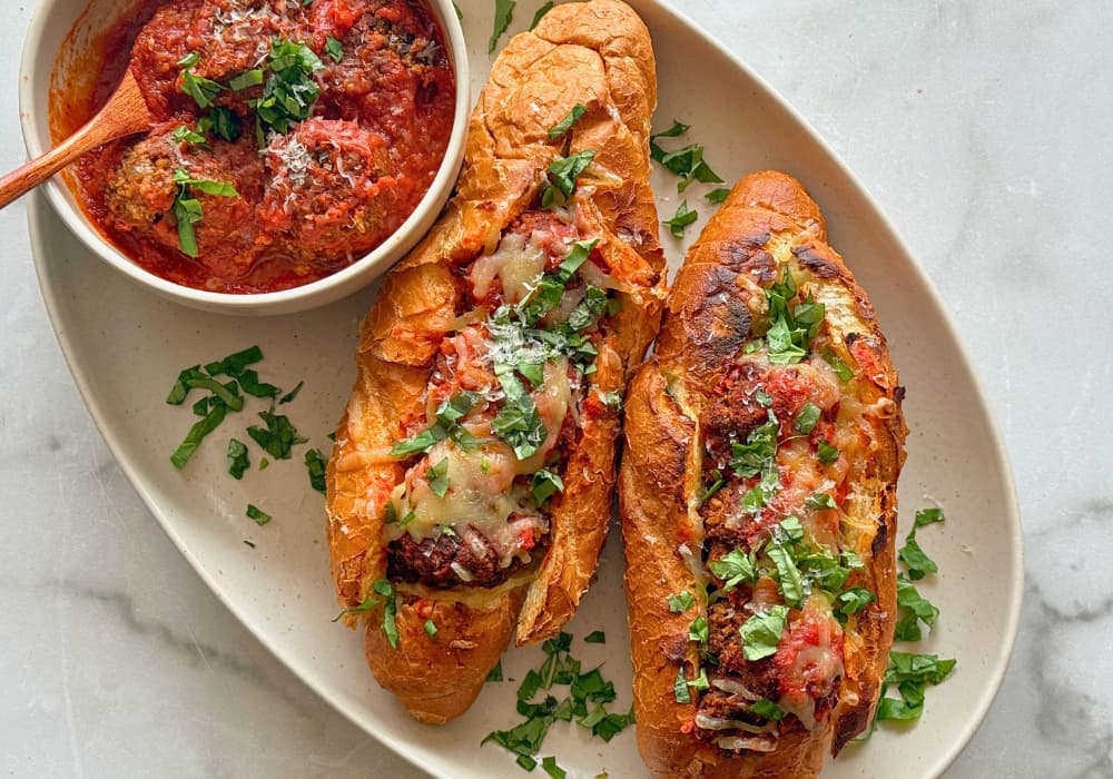 Two Lentil Meatball Subs on an oval platter, filled with lentil meatballs, marinara, and melted dairy-free mozzarella, topped with fresh basil, with a bowl of meatballs in marinara on the side.