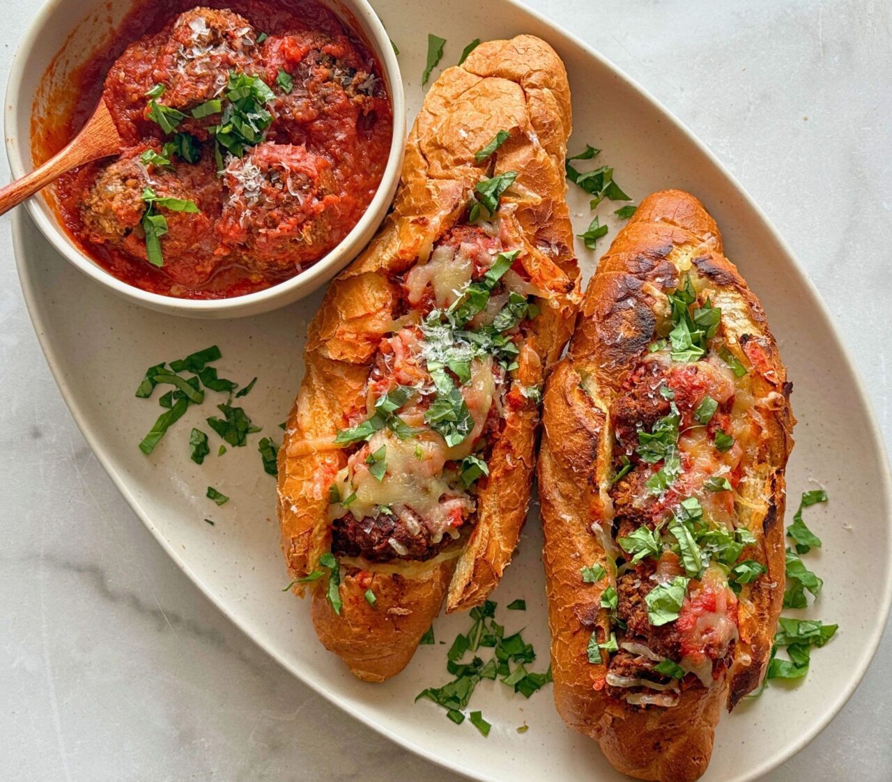 Two Lentil Meatball Subs on an oval platter, filled with lentil meatballs, marinara, and melted dairy-free mozzarella, topped with fresh basil, with a bowl of meatballs in marinara on the side.