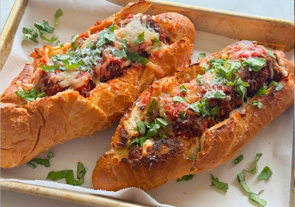 Two Lentil Meatball Subs on a parchment-lined sheet pan, topped with marinara, melted dairy-free mozzarella, and fresh basil.
