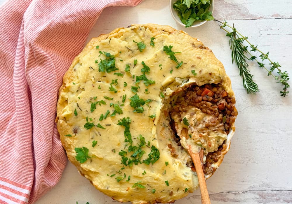 A vegan lentil shepherd’s pie with a golden mashed potato topping and fresh parsley, with a scoop taken out to show the lentil and vegetable filling underneath.