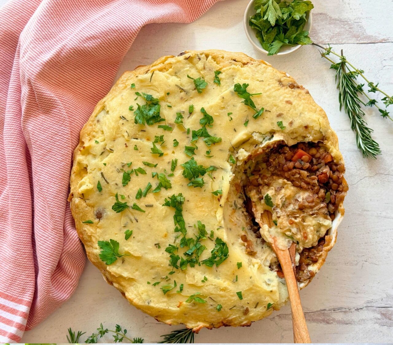 Lentil Shepherd’s Pie with Rosemary Mashed Potatoes with a scoop taken out to show the lentil and vegetable filling, garnished with fresh parsley, rosemary, and thyme.