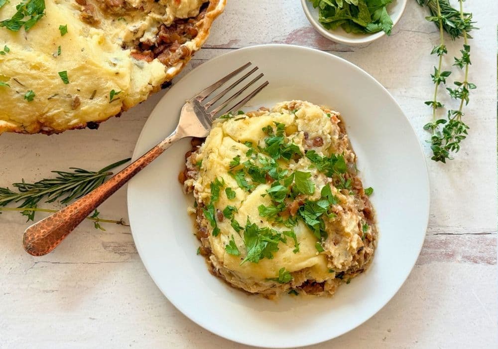 A serving of Lentil Shepherd’s Pie on a white plate, topped with rosemary mashed potatoes and fresh parsley, with a copper fork.