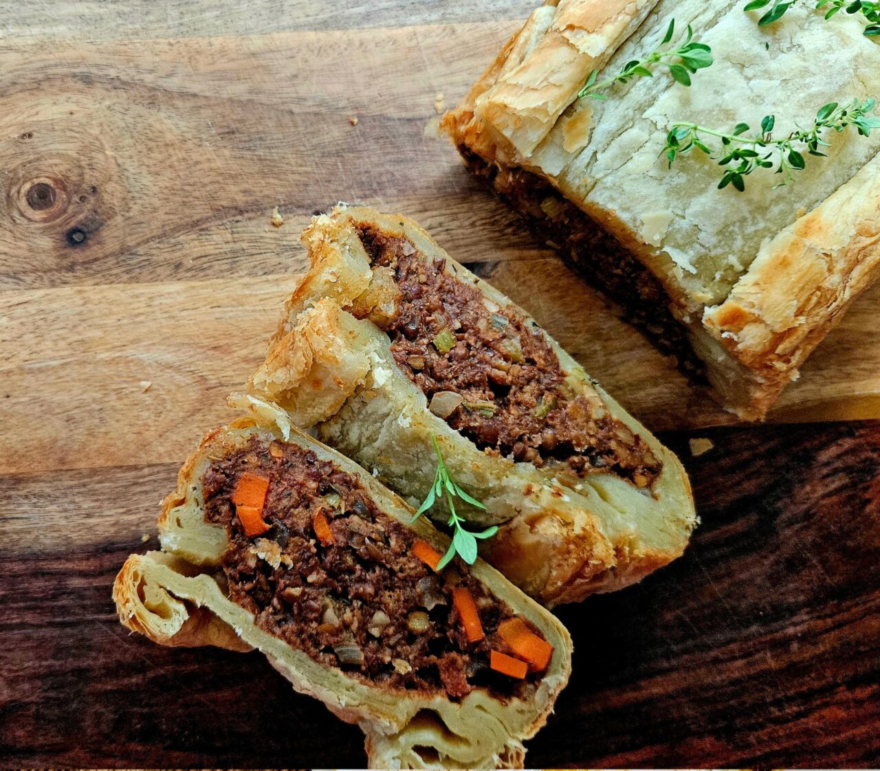 Lentil Wellington on a wooden board with two slices cut to show the lentil, walnut, and carrot filling inside flaky puff pastry, garnished with fresh thyme.