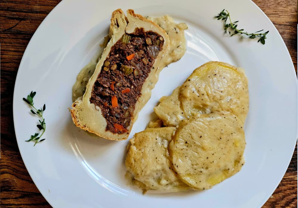 A slice of Lentil Wellington on a white plate with classic gravy and scalloped potatoes, garnished with fresh thyme.