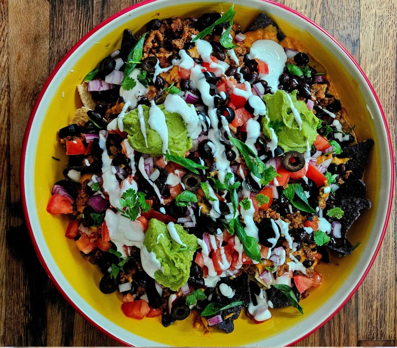Overhead view of Loaded Nachos with Creamy Queso in a yellow bowl, topped with plant-based ground beef, black beans, black olives, tomato, red onion, guacamole, sour cream, and cilantro.