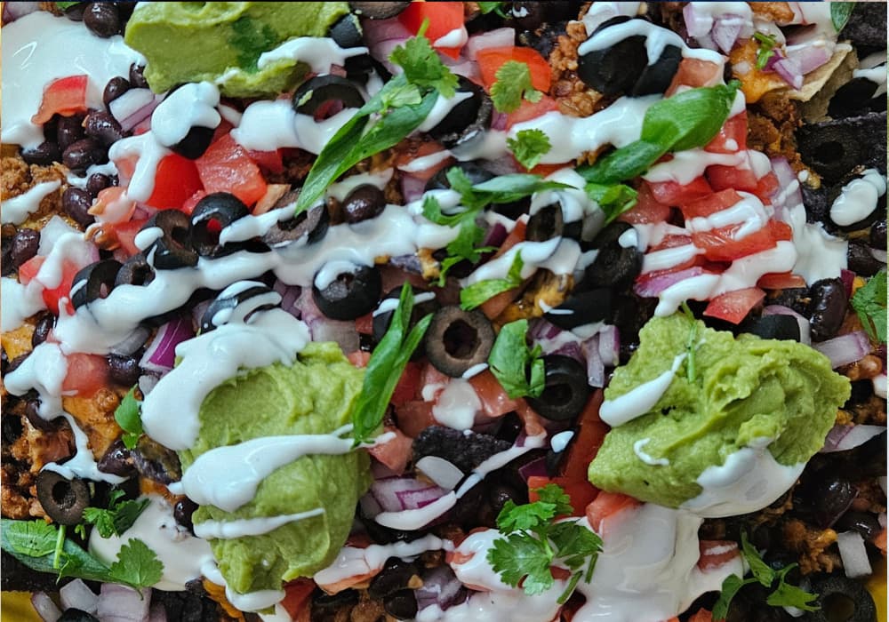 Close-up of Loaded Nachos with Creamy Queso topped with black beans, plant-based ground beef, black olives, diced tomato, red onion, guacamole, sour cream, and fresh cilantro.