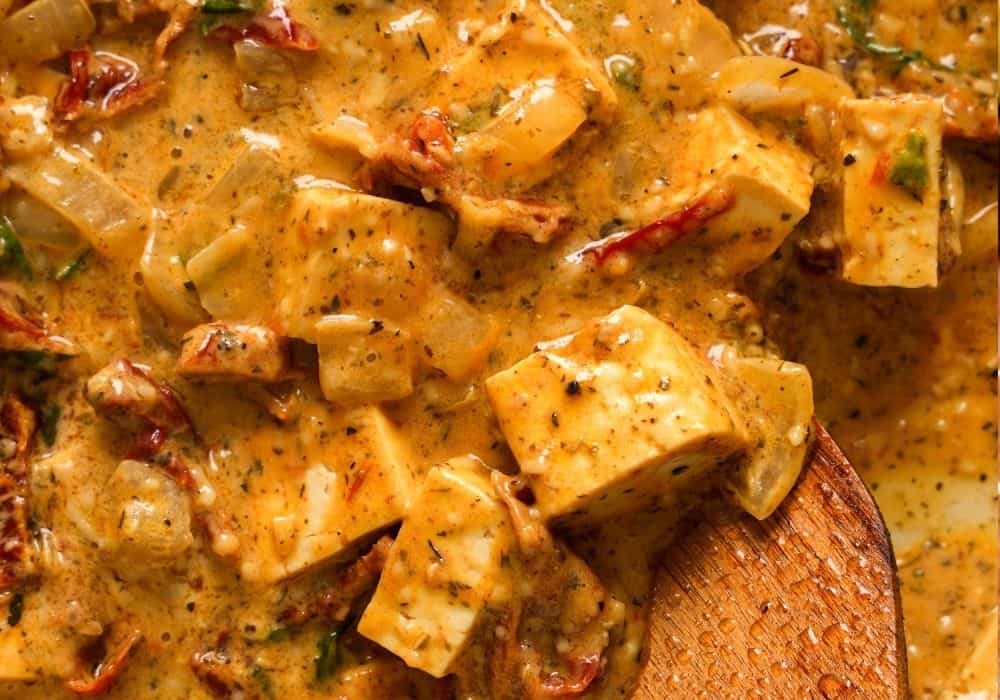 Close-up of creamy Marry Me Tofu with tofu cubes, sun-dried tomatoes, and onion in a dairy-free sauce with a wooden spoon.
