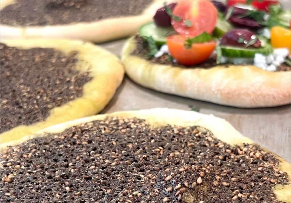 Traditional Mediterranean flatbread with za’atar and olive oil in the foreground, with a flatbread loaded with cherry tomatoes, cucumber, olives, and dairy-free feta behind it.