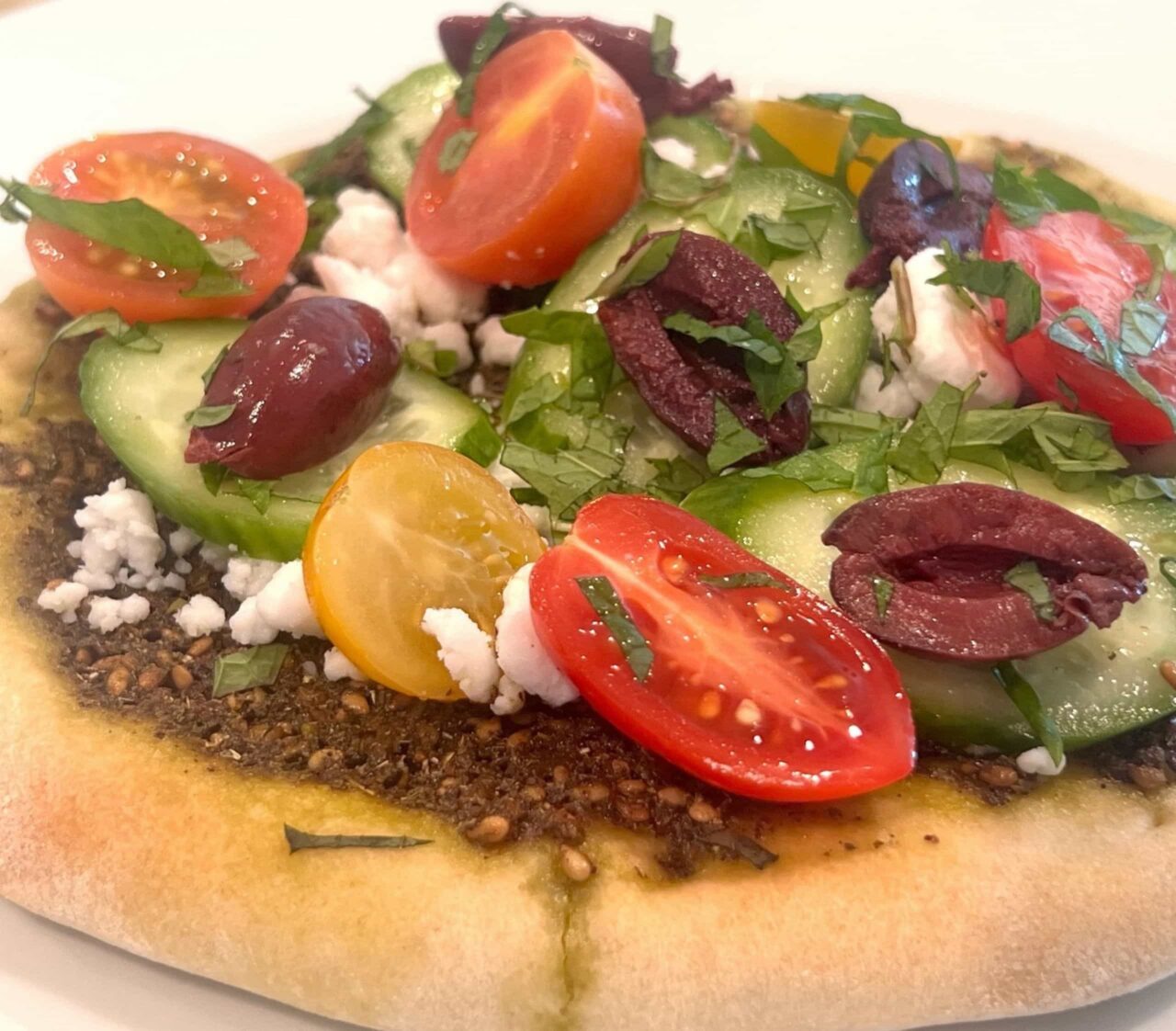 Easy Mediterranean Flatbread on a white plate, topped with za’atar, crumbled dairy-free feta, red and yellow cherry tomatoes, cucumber slices, kalamata olives, and fresh mint.