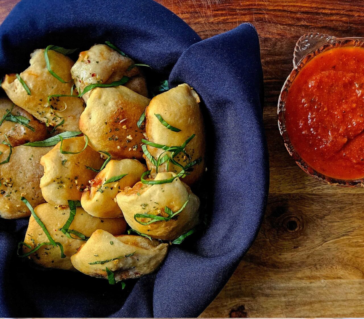 Golden Mini Sausage Pizza Pockets topped with fresh basil and red pepper flakes in a basket lined with a blue napkin, served with a bowl of marinara sauce.