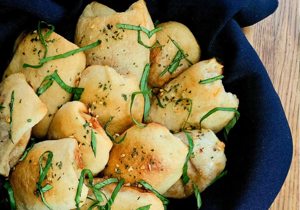 Close-up of golden Mini Sausage Pizza Pockets in a basket lined with a blue napkin, topped with fresh basil, and dried herbs.