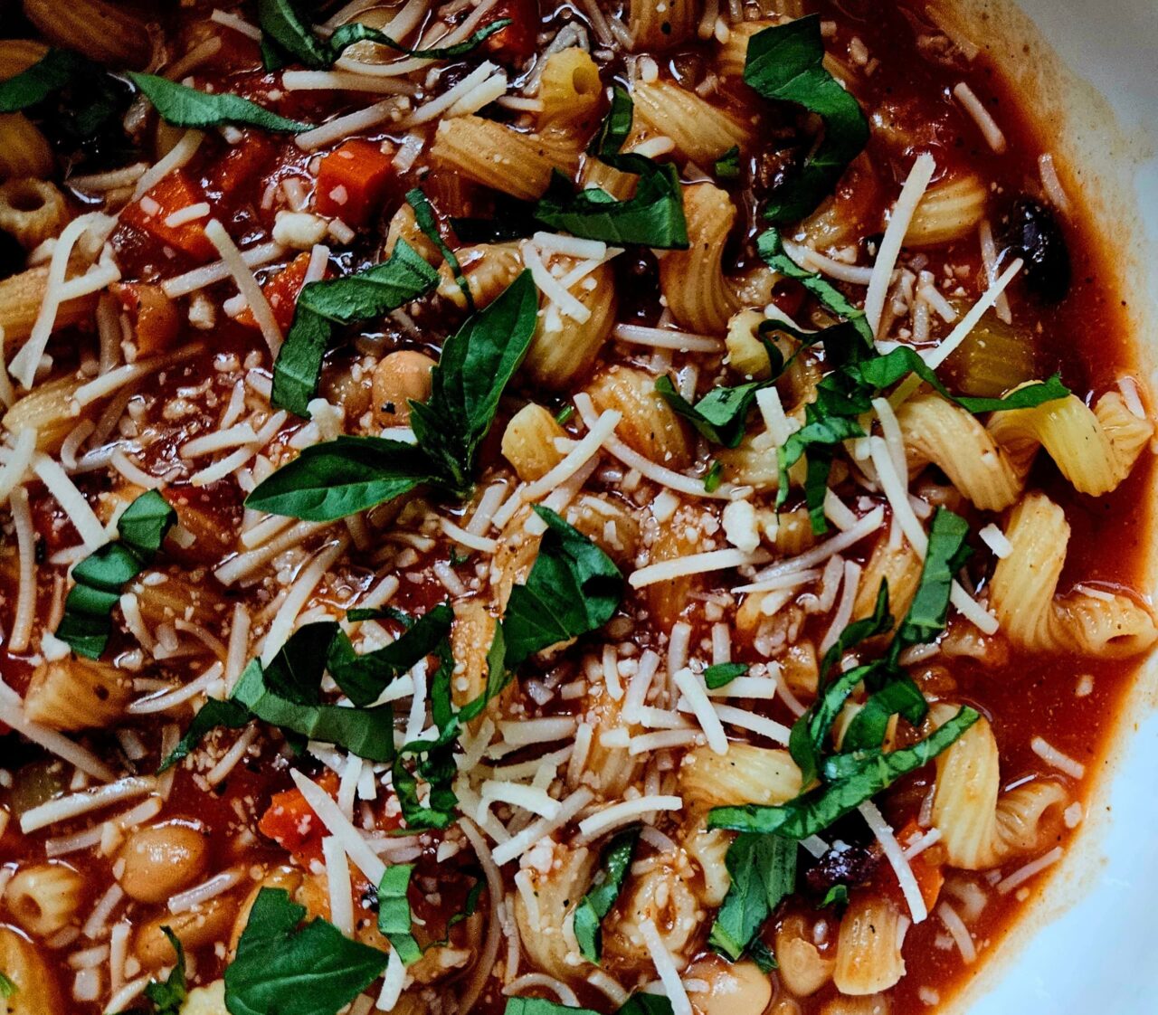 Close-up of Pasta e Fagioli with short pasta, beans, carrots, and celery in a tomato broth, topped with fresh basil and grated parmesan.