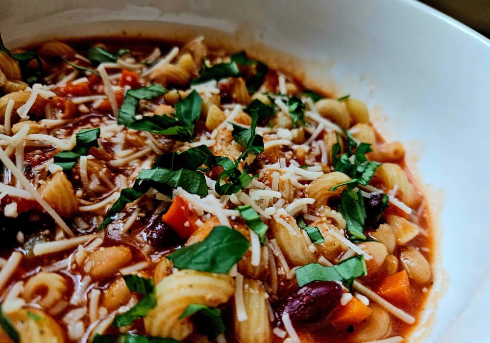 A bowl of Pasta e Fagioli with short pasta, white and kidney beans, carrots, and tomato broth, topped with fresh basil and grated parmesan.