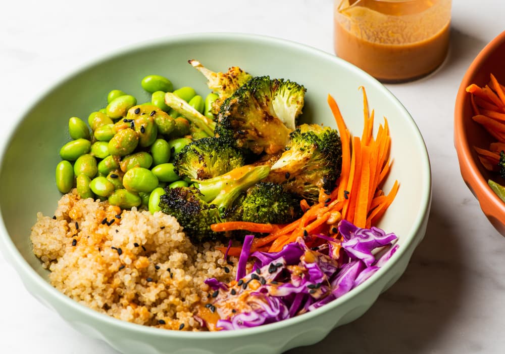 A Buddha bowl filled with quinoa, edamame, roasted broccoli, shredded carrots, and red cabbage drizzled with soy-ginger dressing, with a jar of dressing in the background.