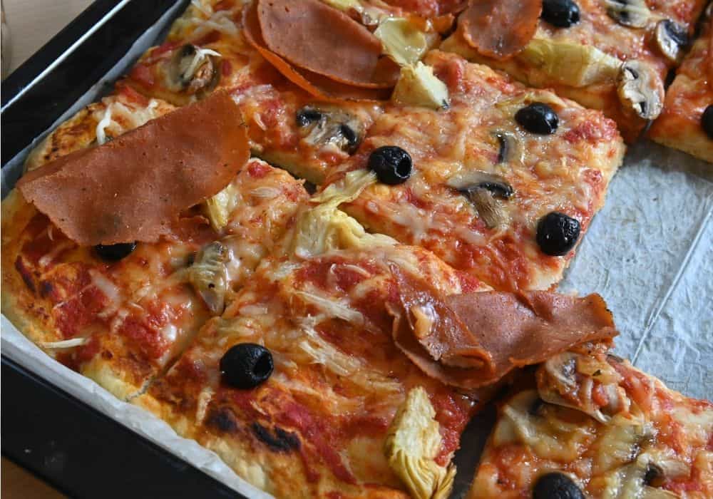 Sheet Pan Pizza with Mushrooms, Artichokes, and Olives cut into squares in a baking sheet, topped with melted dairy-free mozzarella, plant-based ham, and black olives.