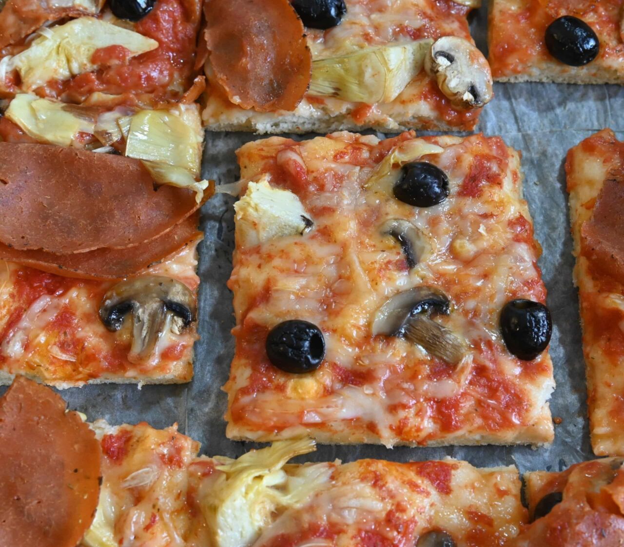 Squares of Sheet Pan Pizza with Mushrooms, Artichokes, and Olives, topped with melted dairy-free mozzarella, tomato sauce, and plant-based ham.