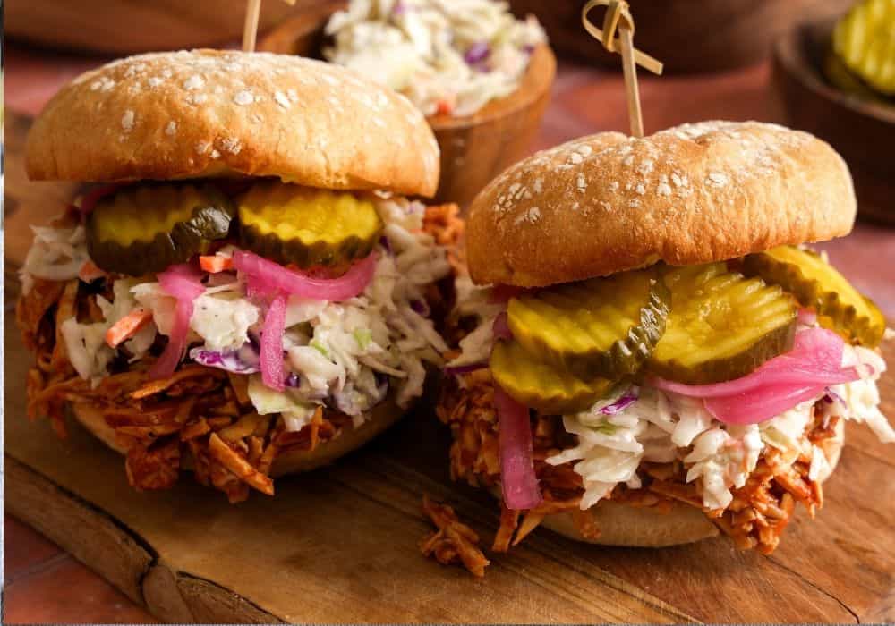 Two shredded BBQ tofu sandwiches topped with creamy coleslaw, pickled red onions, and bread and butter pickles on a wooden board.