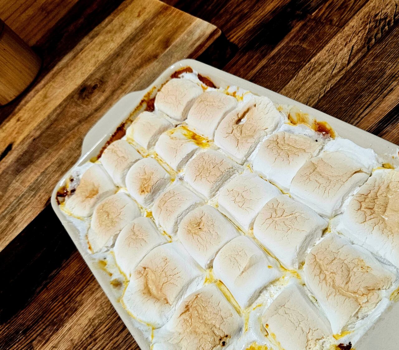 A baking dish of Sweet Potato Casserole topped with rows of golden, toasted marshmallows on a wooden table.