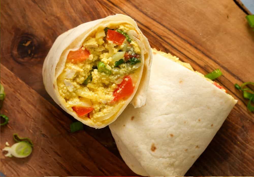 Overhead view of a tofu scramble breakfast burrito sliced in half on a wooden board, filled with tofu scramble, hash browns, red bell pepper, and green onions.