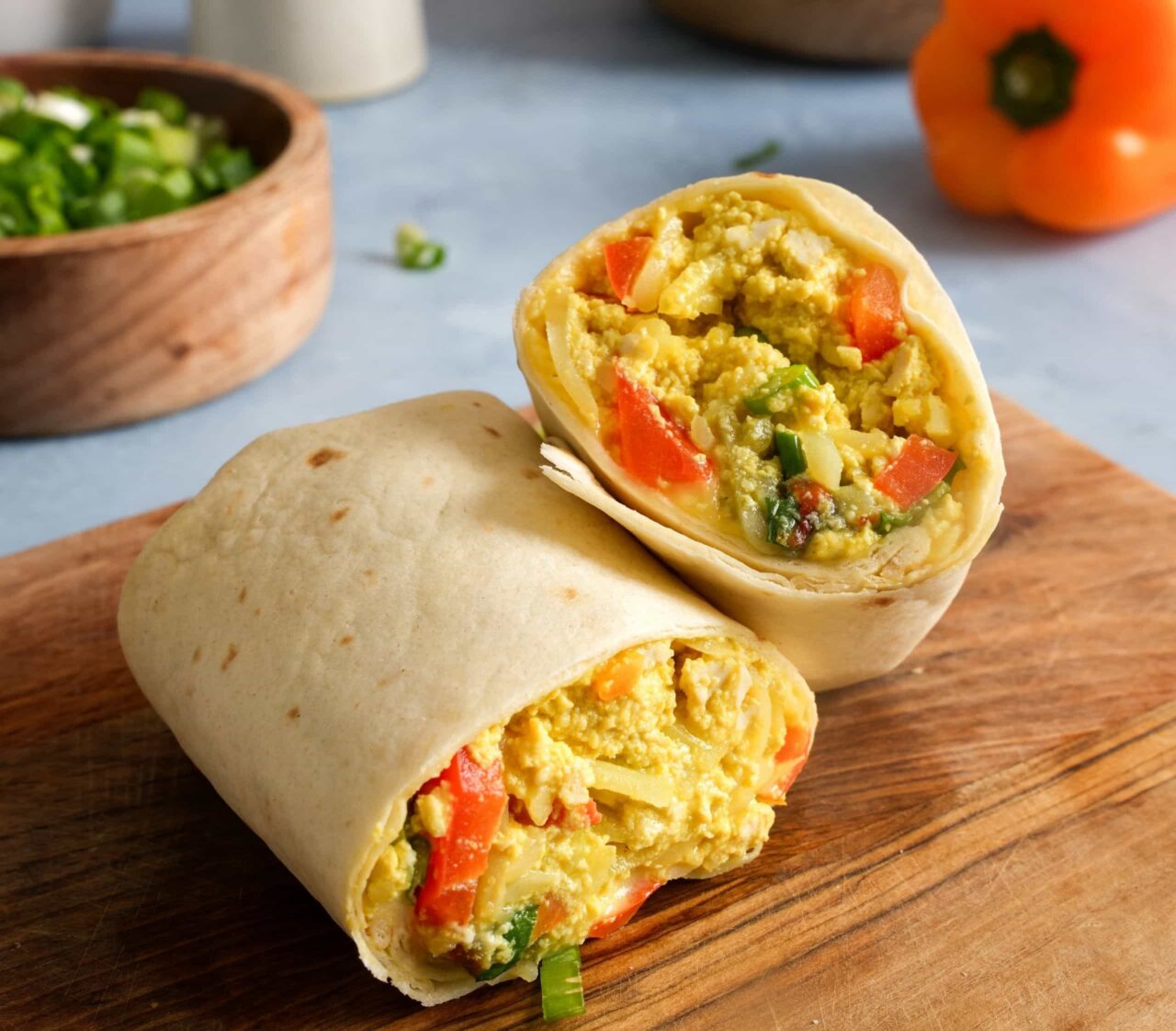 Tofu scramble breakfast burrito cut in half on a wooden board, showing creamy tofu scramble, hash browns, red bell pepper, and green onions.