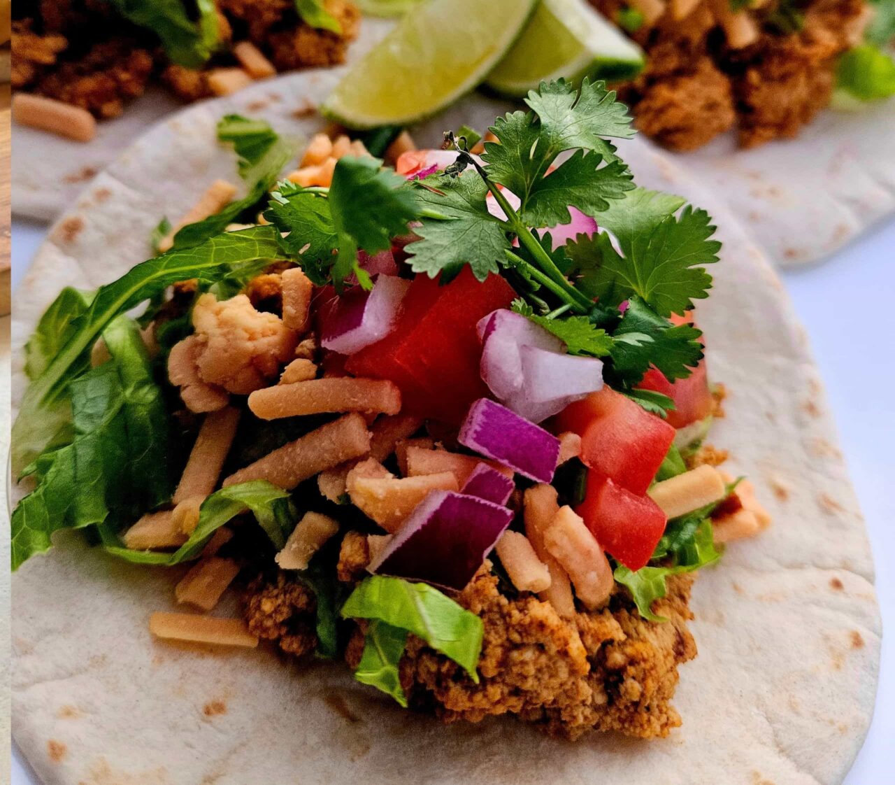 Tofu street taco on a flour tortilla topped with seasoned crumbled tofu, lettuce, shredded cheddar, diced tomato, red onion, and fresh cilantro, with lime wedges.